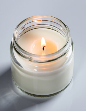 A lit white candle in a clear glass jar, casting a warm glow on a reflective surface.