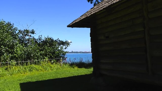 Old traditional and vintage log cabin with a splinter roof or wood chip roofing at the baltic sea blue ocean seaside shoreline beach during a summer sunny day, horison visible besides the trees.