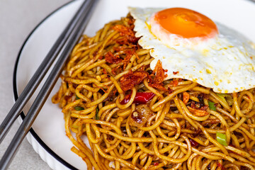 Close-up of homemade fried noodles with a sunny-side-up egg and metal chopsticks served on a white plate.
