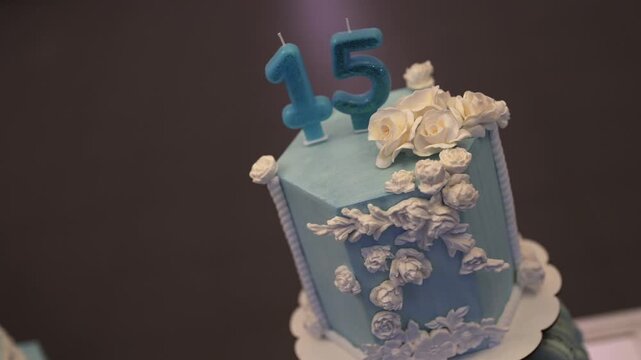 Slow reveal showcases a hexagonal blue fondant cake adorned with cascading white flowers and number candles “15,” highlighting artistic detail and celebration mood for a milestone birthday.