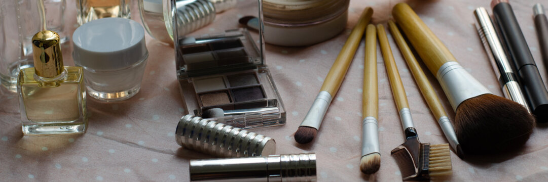 A wide, panoramic shot of a collection of makeup products and brushes, including eyeshadow, perfume, and lipstick, arranged on a light-colored, polka-dotted cloth.
