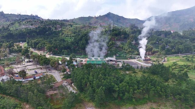 Aerial view of geothermal power plant operating amid lush forested hills, with visible steam vents releasing geothermal heat into the atmosphere. Dieng, Indonesia.
