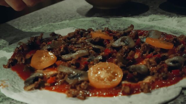 Chefs hand placing and spreading sliced mushrooms and other pizza toping on raw fluffy and hite pizza dought hat has tomato sauce, minced meat and tomato sliced on it. Slow motion movement, bokeh.