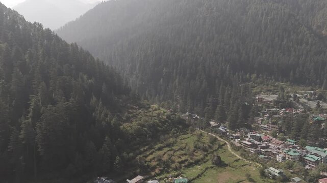 Aerial view of Parvati Valley near Chalal village in Himachal Pradesh, featuring scattered village houses, rocky Himalayan mountains, and dense pine&ndash;deodar forests under a cloudy autumn sky.