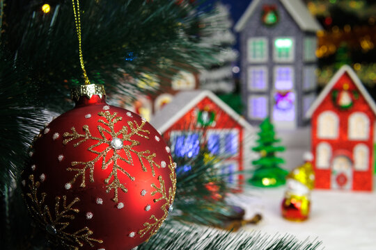 Behind the red ornamental ball hanging on a Christmas tree branch, a Christmas village is partially blurred