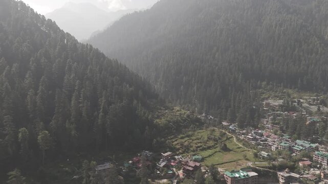 Aerial view of Parvati Valley near Chalal village in Himachal Pradesh, featuring scattered village houses, rocky Himalayan mountains, and dense pine&ndash;deodar forests under a cloudy autumn sky.