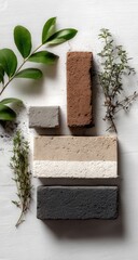 Four rectangular blocks of varying earth-toned colors, arranged vertically, are surrounded by sprigs of greenery and dark soil on a white backdrop.
