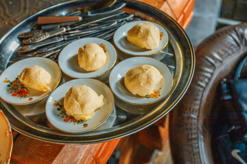 A metal tray holds five small floral-patterned plates, each with a mound of pale dough or butter. Ornate silverware rests in the background, set on a rustic wooden table — a charming, traditional food