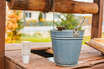  rustic scene: a metal bucket with a handle hangs from a wooden well frame, with water dripping around it. Beside it, a stack of white disposable cups sits on a wooden surface — evoking a countryside 