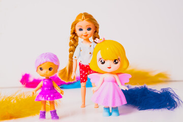Three cheerful toy dolls display vibrant colors and styles. One has long hair with a braid, while the others wear colorful outfits and wings. They are surrounded by playful feathers.