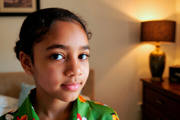 Portrait of teenage girl with medium skin tone looking into camera, standing indoors with soft lighting, showing calm facial expression and subtle smile, hair pulled back