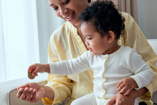 Biracial woman holding toddler, smiling while young child placing coin into her hand, both sitting together near window, mother engaging with baby in playful financial learning moment