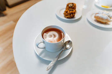 cappuccino with heart latte art on white table, saucer and spoon beside small plates of pastries, soft natural light, clean minimal cafe aesthetic, warm creamy foam, inviting breakfast mood