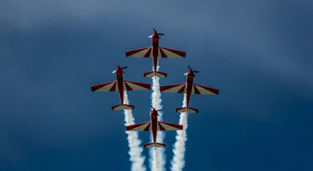 Four acrobatic planes perform a diamond formation in a clear blue sky, trailing white smoke.
