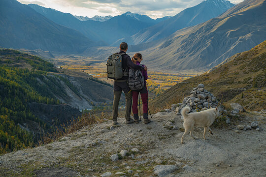 A hiking couple with backpacks embraces on a mountain road edge, admiring the vast autumn valley with peaks, colorful forest, and a blue river. Their large white dog stands nearby.