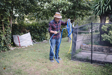 Senior man in hat and flannel winding a blue garden hose with spray nozzle in his green backyard, enjoying simple summer yard maintenance and outdoor retirement chores
