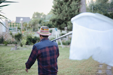 Senior man wearing a plaid shirt and colorful hat carrying a butterfly net over his shoulder, walking through a green backyard, indulging in an entomology hobby