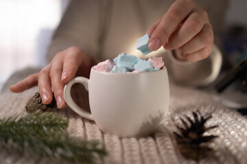 two hands arranging pastel marshmallows above oversized mug, caregiver preparing cozy morning ritual on knit