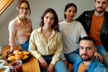 Group of young adult Caucasian and Hispanic men and women sitting together at table with pastries and jam, smiling and looking at camera, casual gathering in modern kitchen