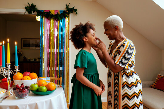 Middle aged Black woman gently touching face of Black girl in green dress while standing together near table with fruit and candles during festive family celebration indoors