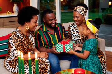 Black man sitting on couch smiling while receiving wrapped gift from Black girl, surrounded by two Black women, all interacting together in living room during family gathering