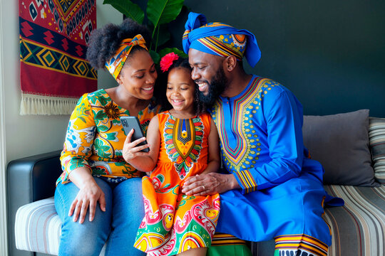 African American family sitting together on sofa smiling and looking at smartphone, middle aged woman, middle aged man, and young girl wearing traditional clothing