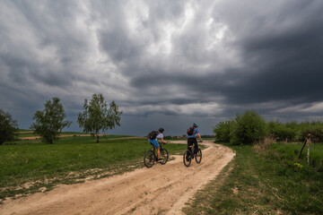 Two cyclists riding on a sandy rural road under heavy storm clouds in the countryside of Małopolska, Poland. Dynamic weather and open fields with scattered trees.
