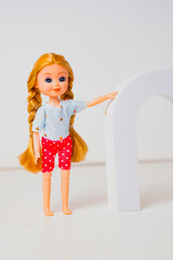 A cheerful toy doll with blonde hair and braided pigtails poses beside a white arch in a bright indoor space. The doll wears a casual outfit with red polka-dotted shorts.
