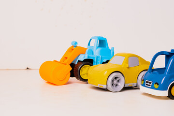 Obraz premium Four colorful toy vehicles are arranged in a row on a light wooden surface. The toys include a yellow truck, a blue car, a green vehicle, and an orange construction machine.