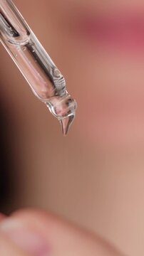 Extreme close-up of clear serum drops slowly falling from a dropper onto fingers, demonstrating precise skincare application and hydration for healthy skin. An unrecognizable woman holds a pipette