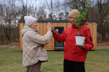 A man in a red jacket and a woman in a quilted coat share a joyful moment as they prepare to plant a small tree in their garden during autumn
