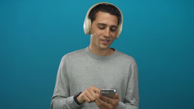 Young hispanic man with headphones using smartphone over isolated blue wall with focus on technology and lifestyle