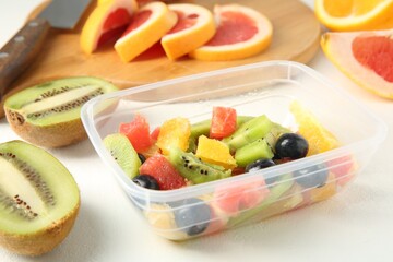 Tasty fruit salad in plastic container, ingredients and knife on white table, closeup. Healthy snack