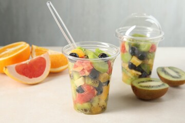 Tasty fruit salad in plastic cups and ingredients on beige table, closeup. Healthy snack