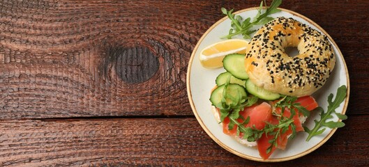 Delicious bagel with salmon, cucumber, arugula and lemon on wooden table, top view. Space for text