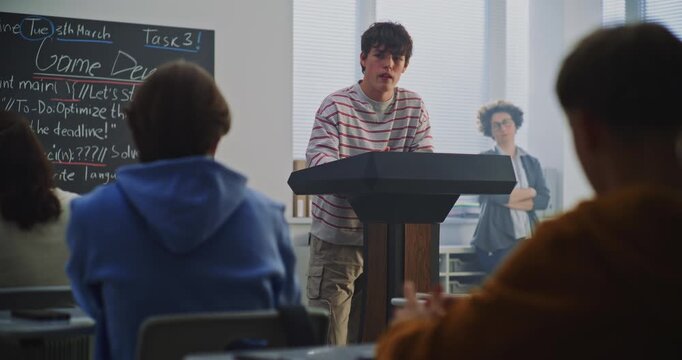 Teenage Boy at Podium Explains Work Peers, Game Dev Topic Visible on Blackboard. Concept Computer Science Education, Process of Peer Review and Discussion in Modern, Technology Focused Classroom.