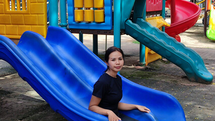 Smiling Descent: A woman takes a moment of pure enjoyment, gliding down a bright blue slide in a vibrant playground, embracing the simple pleasures of play.