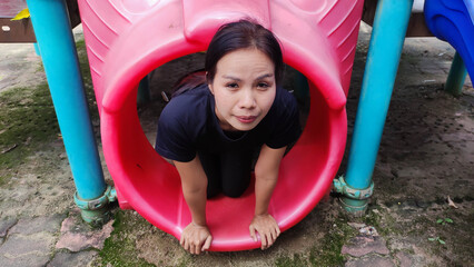Playground Joy: A woman smiles as she peers out from a vibrant red tunnel on a colorful playground, capturing a moment of carefree fun and youthful energy.