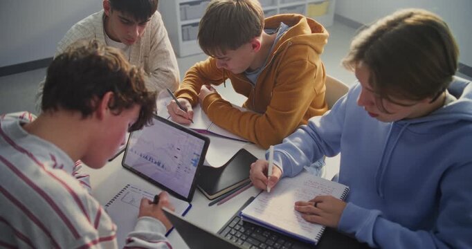Four Young Students Gathers Around Table, Intensely Focused on Collaborative Study Session. Boys Actively Writing In Notebooks and Consulting Digital Tablet Displaying Charts and Graphs. Static Shot.