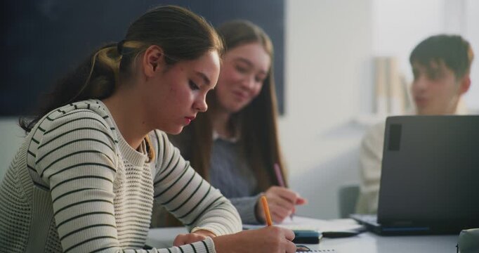 Close Up Two Female Students Working Side by Side at Shared Table. One Girl Writing Attentively in Notebook, Other, in Striped Sweater, Leans in, Looking at Papers. Concept Teamwork, Academic Support.