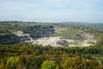 Panoramic View Of An Active Quarry At Black Rocks, Cromford, Derbyshire, Showing Terraced Rock Formations, Autumn Trees, And Distant Countryside Under A Clear Sky.