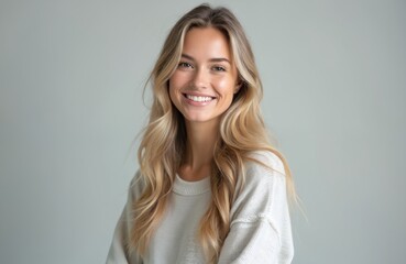 Fototapeta premium Young blonde woman smiles warmly. She wears a light sweater against a plain grey background. Her hair is long and wavy. Pro studio portrait.