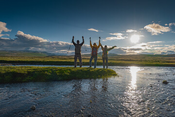 A group of backpackers celebrates on a green grassy island in a mountain river at golden hour. Silhouetted with raised arms against the sunset, with sun reflecting in the water.

