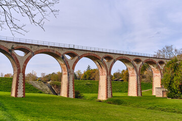 Fototapeta premium Historic brick railway viaduct with stone arches crossing a green valley near a small reservoir in Boulogne-sur-Gesse, symbolizing French industrial heritage and civil engineering 