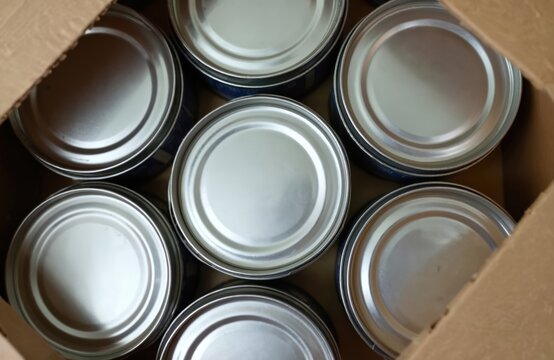 Group of shiny silver cans inside a brown cardboard box. These unopened metal containers are ready for food delivery and storage. They suggest pantry stocking and meal preparation.