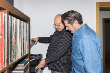Two mature men looking at a turntable as one of them plays a vinyl record. Realistic and warm home scene, horizontal shot with copy space. Concept of friendship and music lifestyle
