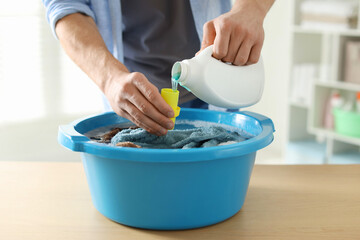 Man pouring detergent into cap over plastic basin with towels at wooden table indoors, closeup