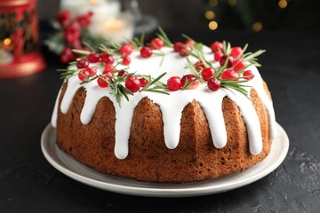 Tasty Christmas cake with icing, cranberries and rosemary on black table against blurred lights, closeup