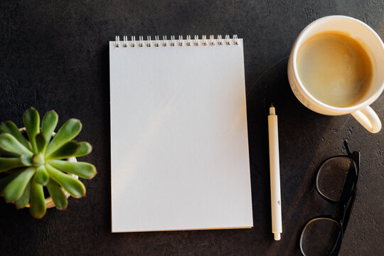 Blank sketchbook with spiral binding, a pen, a cup of coffee, and glasses on a dark surface, creating an inviting workspace atmosphere for creativity and inspiration