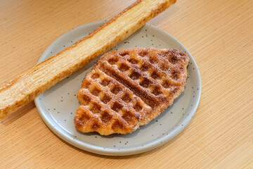 A croffle with cinnamon sugar topping and a slice of garlic bread served on a gray ceramic plate over a wooden table in natural light. Perfect for bakery advertising, cafe menu